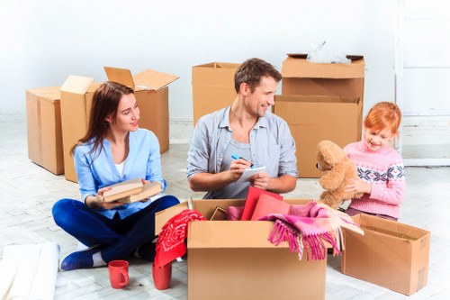 A family of three, including a woman with long brown hair dressed in a blue top and jeans, a man with short dark hair wearing a grey shirt, and a young girl with red hair in a pink sweater, are sitting on a light wooden floor inside a spacious, light-colored room with visible white wall panels. They are surrounded by several open cardboard moving boxes of various sizes, some containing household items like a red scarf, a pink cloth, and a small teddy bear, which the girl is holding. The woman is holding a closed cardboard box, while the man is smiling and holding a pen and notebook, possibly for organizing or packing. In the background, there are additional large closed cardboard boxes stacked against the wall, indicating they are in the process of packing or unpacking. A red mug is placed on the floor near one of the boxes, and the natural lighting highlights the domestic setting typical of house removals in the Putney area, London, near postcode SW15. The overall scene reflects a family engaged in packing or preparing for a move, with a focus on cardboard boxes and household items, typical of a professional removals service like Putney Man with Van.