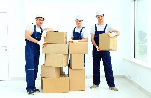 Three professional movers from Putney Man with Van, dressed in blue overalls and white helmets, are standing inside a bright, modern residential space with large windows and white walls. They are surrounded by cardboard boxes of various sizes, some stacked on the floor and others held in their hands, ready for packing or loading. The boxes appear sturdy, sealed with packing tape, and likely contain household items such as clothing, books, or kitchenware. The outer atmosphere is well-lit with natural daylight streaming in through the windows, highlighting the clean, minimalist interior. The movers are smiling and positioned in a manner that suggests they are preparing to transport the boxes, reflecting a professional approach to house removals. The setting emphasizes a typical domestic environment in the London borough of Putney, with a focus on efficient packing and loading activities characteristic of local residential moves. This scene exemplifies the careful handling and organized approach typical of reliable removals services in South West London, as provided by Putney Man with Van.