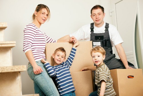 A family of four, including a woman in a red and white striped long-sleeve shirt and two young boys dressed in striped shirts, are helping to move boxes into a home, assisted by a male removal specialist wearing a white shirt and black work uniform. They are positioned in a well-lit hallway near a staircase with wooden steps, and various cardboard boxes of different sizes are stacked around them, some being held or carried. The scene captures a moment of collaborative effort during a house relocation, with the family smiling and engaged in the process of packing or unpacking. The background shows a white wall and a partly visible doorframe, suggesting a residential property in a suburban area, possibly in the Putney or south-west London district, consistent with the company's local service area. The environment indicates a typical domestic setting with natural lighting highlighting the furniture and personal belongings, emphasizing the practical, professional nature of house removal services provided by Putney Man with Van.