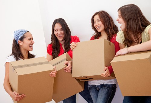 Four women, dressed casually in a brightly lit interior space, are smiling and engaged in conversation while holding large cardboard boxes. The woman on the left, wearing a blue headscarf and white top, is holding a box with both hands, her expression cheerful. The second woman from the left, in a red top with long dark hair, is also holding a box and appears to be speaking to the others. The third, with curly brown hair and a white top, is smiling broadly and unboxing a medium-sized box. The woman on the right, wearing a yellow top, is leaning on a box with her forearm, sharing a moment of conversation. The background suggests a modern residential setting, possibly a hallway or living room, with plain white walls. Various household items, such as a cardboard box and the women's casual clothing, indicate they are preparing for a house move or relocation in the Putney area, part of a professional removals service by Putney Man with Van. The scene captures the process of packing and organizing belongings for a residential move, emphasizing the friendly and efficient nature of the service. Bright, natural lighting highlights the textures of cardboard, textiles, and the women’s hair, creating an inviting and professional atmosphere related to household removals in London.