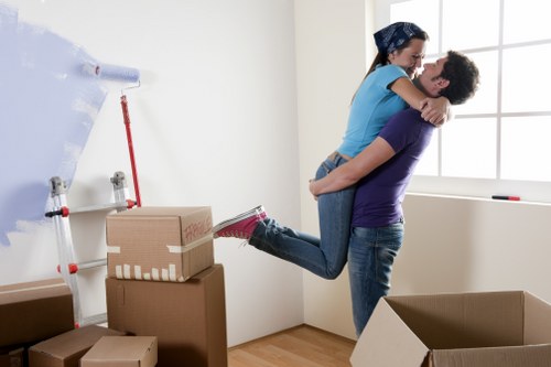 A young couple engaged in a joyful moment during house moving; the man, wearing a purple T-shirt and jeans, is lifting the woman, who is dressed in a blue shirt, dark jeans, and sneakers, off the ground inside a bright, modern room with white walls and a large window. The woman has her arms around the man's shoulders as they smile at each other. Surrounding them are several cardboard boxes, some sealed with tape, placed on the wooden floor, indicating they are in the process of packing or unpacking. In the background, a partially unrolled roll of bubble wrap and a red-handled tool leaning against the wall are visible, along with additional packing materials. The scene suggests an ongoing residential move in a house, possibly within or near Putney, with natural daylight illuminating the space, creating a warm, cheerful atmosphere typical of house removals and relocation activities by Putney Man with Van, emphasizing their professional local service.