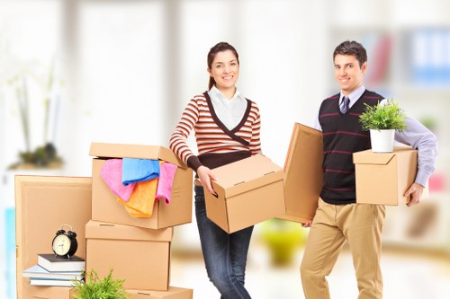 A man and woman, both smiling and dressed casually, are standing indoors in a bright, modern living space with large windows allowing natural light. They are surrounded by several packed cardboard boxes of various sizes, some sealed with tape, others open showing contents like folded towels and books. The woman, positioned on the left, is holding an open box with her left hand while her right hand rests on a stack of closed boxes with colorful towels on top. The man, on the right, is holding a medium-sized box in his right hand and a smaller box with a potted green plant in his left, both boxes appearing ready for moving. The background features a light-colored, minimally decorated room with soft focus, emphasizing the household items and the team’s preparation for a move within London or nearby areas, reflecting services offered by Putney Man with Van in the house removals sector. The scene captures a moment of packing or arranging belongings, illustrating a professional and organized approach to house removals and relocation services.