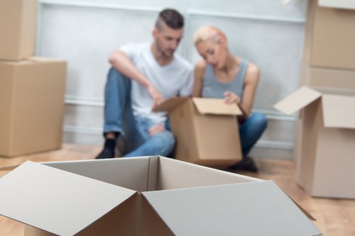 A young man with dark hair and a beard, wearing a white t-shirt and jeans, and a blonde woman wearing a light grey sleeveless top, sit on the floor inside a residential property surrounded by stacked cardboard boxes of various sizes, some opened and partially packed, in a room with white walls and wooden flooring. They are engaged in packing or unpacking belongings, focusing on a medium-sized box in front of them. The scene shows a typical house removal process, with several closed boxes nearby and an open box in the foreground, all prepared for a move within a postcode area that could be associated with Putney, London, emphasizing the local context. The environment appears bright with natural light, suggesting daytime, and the overall setting is indicative of a house moving or relocation service in the area, provided by Putney Man with Van.