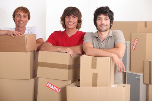 The image features three young men with light skin smiling and standing behind a collection of cardboard boxes, some of which are labeled 'Fragile,' in what appears to be a residential interior or moving environment. The man on the left is wearing a white t-shirt, the one in the middle has shoulder-length brown hair and is dressed in a red shirt, while the man on the right has dark wavy hair and is wearing a light grey t-shirt. They are positioned closely together, with their arms resting on the boxes, suggesting a friendly, collaborative atmosphere typical of house removals or moving services. The background includes a plain white wall and a glimpse of a light-colored piece of furniture or possibly kitchen cabinetry, indicating an indoor setting. The boxes vary in size, some stacked and others placed side by side, with a mix of plain and taped packaging, suitable for relocating household belongings within a local area such as Putney or nearby London neighborhoods. The overall scene emphasizes organized packing and teamwork involved in home removals, highlighting the professional services offered by Putney Man with Van.