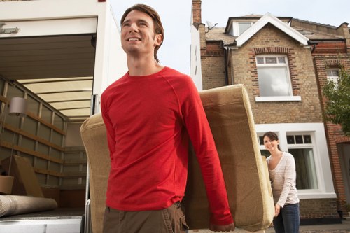 A young man wearing a red long-sleeved shirt and brown trousers is outside a residential property, carrying a large beige upholstered armchair on his shoulder. Behind him, a woman dressed in a light-colored top and dark trousers is standing near an open moving van, ready to assist with the move. The background features a brick house with white-framed windows and a driveway, indicating a suburban setting possibly within Putney or nearby London postcode. The scene is illuminated by natural daylight, with visible elements such as the van's rear cargo area, some cardboard boxes, and household furniture, suggesting an ongoing house removal or relocation process typical of professional removals services.