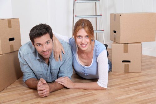 A man and woman lying on a light wooden floor inside a house, surrounded by several cardboard moving boxes, some stacked and others on the ground. The man, with dark hair and wearing a light blue denim shirt, is positioned on the left, resting on his forearms and smiling at the camera. The woman, with auburn hair tied back and dressed in a blue and white long-sleeve shirt, is on the right, supporting herself on her elbows, also smiling. Behind them, a silver step ladder leans against a white wall, suggesting a house interior or a room being prepared for a move. The scene appears well-lit with natural light, indicating daytime, and the environment reflects a typical residential setting in the UK, with the focus on the process of packing or unpacking as part of a house removal or relocation service provided by Putney Man with Van.