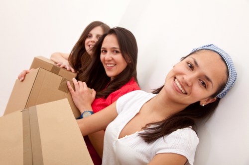 Three women involved in a house removal task are seen in an indoor setting with a plain white wall background. The woman closest to the camera is wearing a white t-shirt and a blue patterned headscarf, smiling while carrying a medium-sized cardboard box with brown tape visible along the edges. Behind her, the second woman, dressed in a red top, is also holding onto the same box, smiling with her dark hair loose. The third woman, further back, is wearing a dark top and has long brown hair, smiling as she assists with additional packaged items. The environment appears to be a residential interior, likely a hallway or living area, with no furniture or other objects visible aside from the boxes. The natural lighting highlights the friendly and collaborative atmosphere typical of house removals in the Putney area, London, suggesting a professional and organized move handled by Putney Man with Van, a local removals service specializing in residential relocations.