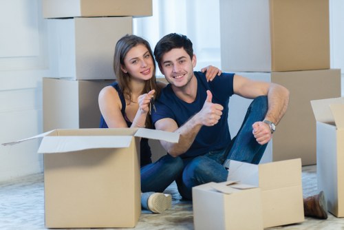 A young couple sits on the floor of a house interior surrounded by open cardboard moving boxes, some filled with household items. The woman has long brown hair and is wearing a dark sleeveless top, while the man has short dark hair and is dressed in a T-shirt and jeans. They are smiling at the camera, with the man giving a thumbs-up gesture. The background shows a bright room with large windows and white curtains, filled with packed boxes of various sizes, some stacked on top of each other. The scene depicts a recent move or packing process, typical of house removals in the Putney area, with the couple appearing happy and relaxed during the packing stage of their relocation. The setting emphasizes an organized and domestic environment, with natural lighting highlighting the textures of the cardboard boxes and the casual home atmosphere.