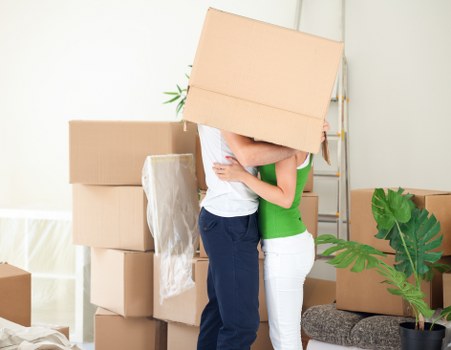 A scene inside a residential interior shows two individuals engaged in a house move, with one person holding a large cardboard box resting on their shoulder while the other supports it from below. The person on the left wears navy blue trousers and a white top, and the person on the right has white trousers and a bright green top. Surrounding them are multiple cardboard boxes of varying sizes, some stacked on the floor and others arranged along the wall, indicating an ongoing packing process. The background includes a light-colored wall, a window that allows natural daylight to brighten the room, and a potted plant with broad green leaves placed on a small table or stool. A grey upholstered chair is visible near the boxes, suggesting a lived-in, recently packed space typical of a house on a suburban street in the Putney area. The scene captures an organized, professional approach by Putney Man with Van to house removals, emphasizing careful handling of household items in a domestic environment, with attention to safety and efficiency during the move.