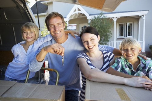 A family of four, including a smiling father with short brown hair, a mother with dark hair, and two children with blonde hair, are outdoors in front of a white residential house with a porch. The father is holding a set of keys in his right hand, while the mother and children are smiling and leaning on cardboard boxes and a trolley with packing materials. The scene suggests a house move, with the family standing behind an open vehicle trunk filled with boxes and household items prepared for transportation. The background shows the house's exterior with windows, a porch roof, and garden greenery, indicating a suburban setting. The lighting is natural daylight, highlighting the cheerful atmosphere of completion or preparation for a move, related to removals services that Putney Man with Van might offer in the local area near Putney or surrounding parts of London.