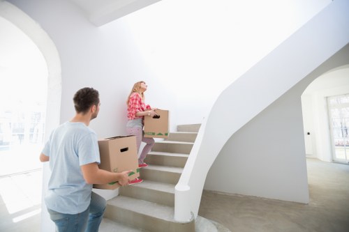 A young woman with blonde hair, dressed in a red checkered shirt and jeans, is carrying a large cardboard box up a staircase in a bright, modern residential interior. A man with dark hair, wearing a light blue t-shirt, is standing at the bottom of the staircase holding a similar cardboard box. The staircase features white walls and a smooth curved handrail, and natural light floods the area through large windows on the left side, illuminating the grey carpeted floor. The background shows an open, airy space with contemporary architectural details, suggestive of a house in the Putney area. This scene illustrates house removals activity, with household furniture and personal belongings being moved during a residence change, supporting a professional removals service in London postcode SW15.