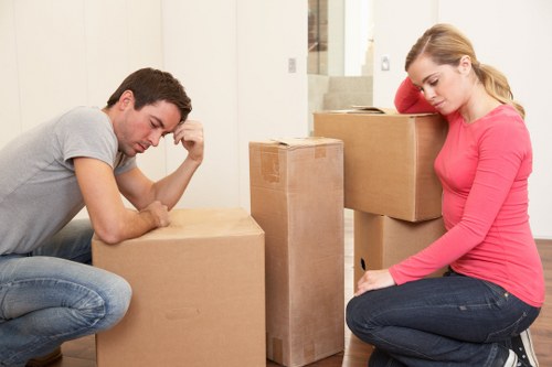A man and a woman are kneeling indoors amidst a collection of packed cardboard boxes, likely preparing for a house move in a residential area near Putney. The man, dressed in a light grey t-shirt and jeans, appears to be deep in thought, with his hand resting on his forehead as he looks at a large sealed cardboard box on the floor. The woman, wearing a bright pink long-sleeve top and dark jeans, is sitting on the floor beside a tall, narrow box, with her eyes downcast and hands resting on her thighs. Behind them, additional larger cardboard boxes are stacked, and the environment looks to be a bright, modern house with neutral-colored walls and a staircase visible in the background. The scene captures a moment of packing or planning during a home removal, with the focus on the packing process typical of professional removals services in the Putney area, emphasizing the careful handling and preparation of household items for a safe move.