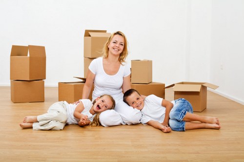 A smiling woman with light blonde hair, wearing a white t-shirt, sitting on a light wooden floor in a bright, empty room, surrounded by numerous cardboard moving boxes of varying sizes, some open and some closed. She is positioned behind two children lying on the floor; a young girl with long blonde hair, dressed in a white top and light-colored pants, lying on her side with her head resting on her arm, and a young boy with short dark hair, wearing a white shirt and blue shorts, lying on his stomach with his arms crossed. The boxes are stacked and arranged around them, indicating a house move in progress, possibly in the Putney area near London, as suggested by the context of moving services. The room has white walls and a clean, minimalist appearance, with no furniture visible. The natural lighting illuminates the scene evenly, emphasizing the cheerful atmosphere and the process of packing or unpacking during a house relocation involving Putney Man with Van or similar removals services.