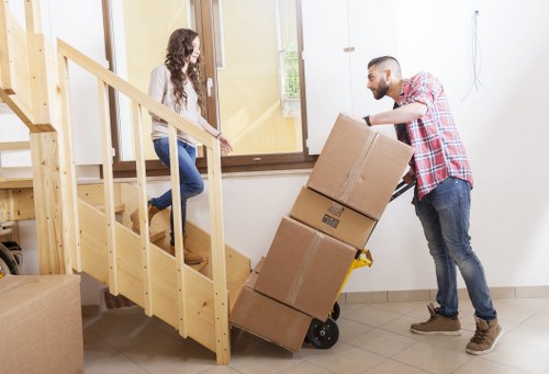 A man and a woman are engaged in a house removal process inside a residential property. The man, wearing a red and white checked shirt and dark jeans, is standing on the floor and guiding a hand truck loaded with three cardboard boxes, stacked one on top of the other. The woman, dressed in a white top and blue jeans, is kneeling on a staircase with wooden steps and a light wood handrail, assisting by handing or receiving items. The backdrop features a cream-colored wall and a large window that allows natural light into the interior, illuminating the tiled floor and part of a white door frame. Nearby, a portion of the staircase is visible, indicating this is an upper-floor or entrance area of a house in a suburban setting. The scene captures a professional removals operation by Putney Man with Van, demonstrating careful handling and collaboration during household furniture and box transportation, with a focus on residential house relocation within the town or postcode area.