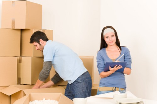 A man dressed in casual clothing, with a light blue T-shirt and jeans, is seen bent over, carefully handling cardboard boxes in a room with white walls, possibly preparing items for a house move within the Putney area. The man is placing or adjusting boxes that vary in size, with several stacked against the wall, some sealed with brown packing tape. A woman, wearing a light blue long-sleeved top, beige trousers, and a white headband, is standing nearby holding a clipboard or notepad, looking at the camera with a slight smile, representing a professional house removals service. In the foreground on a white surface, there are more unpacked or packed boxes, along with white bowls or cups, suggesting a home environment during packing or unpacking. The setting appears to be a residential interior, possibly a living room or hallway, featuring minimal decor and a clean, well-lit atmosphere. The scene reflects the process of residential relocation, emphasizing careful handling of belongings and organized packing, characteristic of a Putney-based removals company.