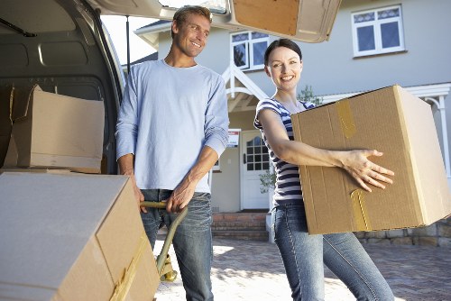 A man and a woman, both smiling, are engaged in house removals outdoors in front of a residential property with a white exterior and large windows. The man, dressed in a light blue long-sleeve shirt and jeans, is standing beside an open removals van, holding a dolly with a cardboard box on it. The woman, wearing a striped t-shirt and jeans, is carrying a large cardboard box with yellow packing tape, holding it securely with both hands. The van’s interior shows additional cardboard boxes stacked neatly inside, ready for loading or unloading. The scene is set on a paved driveway, with the house's entrance visible behind them, and the natural daylight highlights the details of the furniture, boxes, and their casual attire. The overall atmosphere suggests careful preparation for a residential move within the Putney area, demonstrating the professional services provided by Putney Man with Van for house removals and relocations.