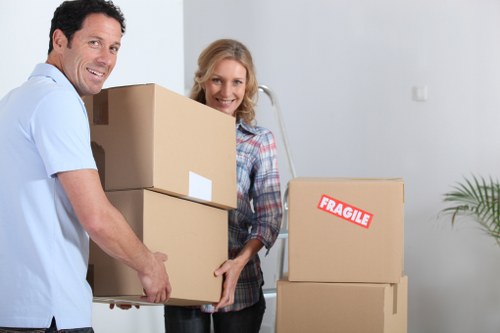 A man and woman involved in domestic relocation, with the man carrying a stack of two cardboard moving boxes, one of which is plain and the other with a white label, while the woman stands nearby, smiling, in a bright, modern interior space with light-colored walls. Several additional cardboard boxes are stacked to the right, one marked with a red 'FRAGILE' label, indicating careful handling. The background includes a small potted plant on the right side and a visible household wall socket, suggesting a residential setting, likely within the Putney area. The scene captures the process of house removals, with both individuals dressed casually and engaged in the moving task, emphasizing professional services by Putney Man with Van. The environment appears well-lit, with natural or ambient lighting highlighting the clean, organized space prepared for a residence transfer, enhancing the visual understanding of a typical house removal operation in London suburbs.