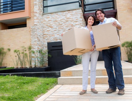 A smiling young couple stands outside a modern residential building on a sunny day, each holding a large cardboard box. The woman, with shoulder-length dark hair, wears a light purple blouse and white trousers, while the man, with short dark hair and glasses, is dressed in a white shirt and dark trousers. In the background, the house has a brick facade with a partial balcony and large windows, suggesting a contemporary style. They are positioned on a paved pathway leading to the entrance, with a well-maintained lawn and small plants on the left side, indicating a suburban neighbourhood. The scene appears to capture a moment of unpacking or moving, with the couple prepared to load or unload belongings, fitting with the services of Putney Man with Van that specialises in house removals and relocations within the local area around Putney, SW London. The bright, natural lighting highlights the domestic setting and the casual, friendly atmosphere of a house move in progress.