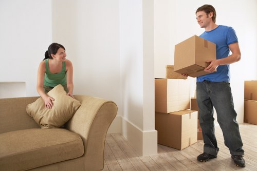 In a bright, minimalist living room with white walls and light wooden flooring, a woman with dark hair tied back, wearing a green sleeveless top and sitting on a beige fabric sofa, is smiling and holding a beige cushion. She appears relaxed and engaged as she interacts with a man standing nearby. The man, with short brown hair and wearing a blue T-shirt and dark jeans, is smiling and holding a medium-sized cardboard box. In the background, there are several larger cardboard boxes stacked along the wall, suggesting a house move or relocation process. The scene is well-lit with natural light, highlighting the clean and organised interior, typical of a residential property in the Putney area, and reflects the work of a professional removals service such as Putney Man with Van, experienced in house removals with attention to customer care and efficiency.