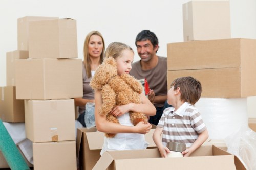 A family of four, including two children and two adults, are engaged in packing up a home for a move in a residential property, with cardboard boxes of various sizes stacked around them. The children, a girl with curly blonde hair wearing a beige furry jacket and a boy with short brown hair wearing a striped shirt, are inside an open cardboard box, handling small items. The girl holds a teddy bear, while the boy is holding a small metallic object. The adults, a woman with long blonde hair and a man with dark hair, are smiling and standing behind the children, observing the activity. The scene is illuminated by natural light, with a plain white wall in the background, indicating an indoor setting such as a living room or hallway in a house in Putney, SW15 postcode. The overall environment suggests a house removal process facilitated by Putney Man with Van, with household furniture and personal belongings being organized for transport, capturing the typical activities involved in local removals service.